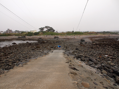 [There are several overhead wires from the mainland to the island sloping upward above the top of the photo. The paved ramp has rocks on either side and rocks from the end of the ramp to the mainland. Two people at the very bottom of the ramp are only visible because their brightly colored clothing constrasts with the brown and black of the rocks and pavement.]